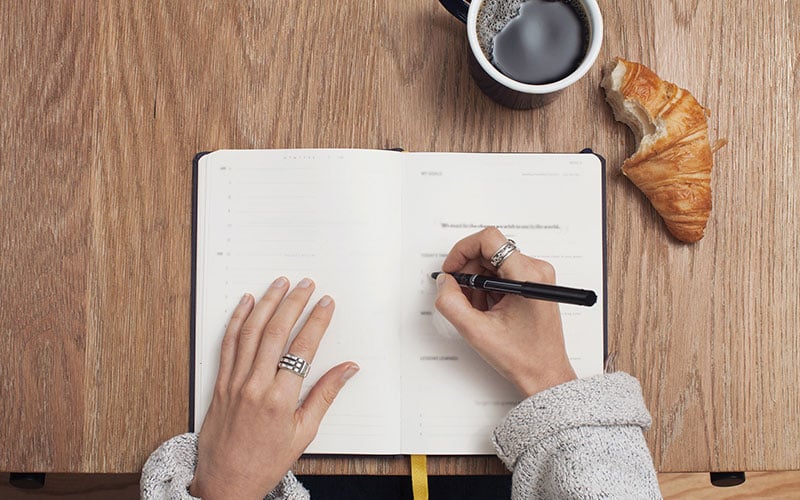 A woman writing in a notebook with a pen and croissant.